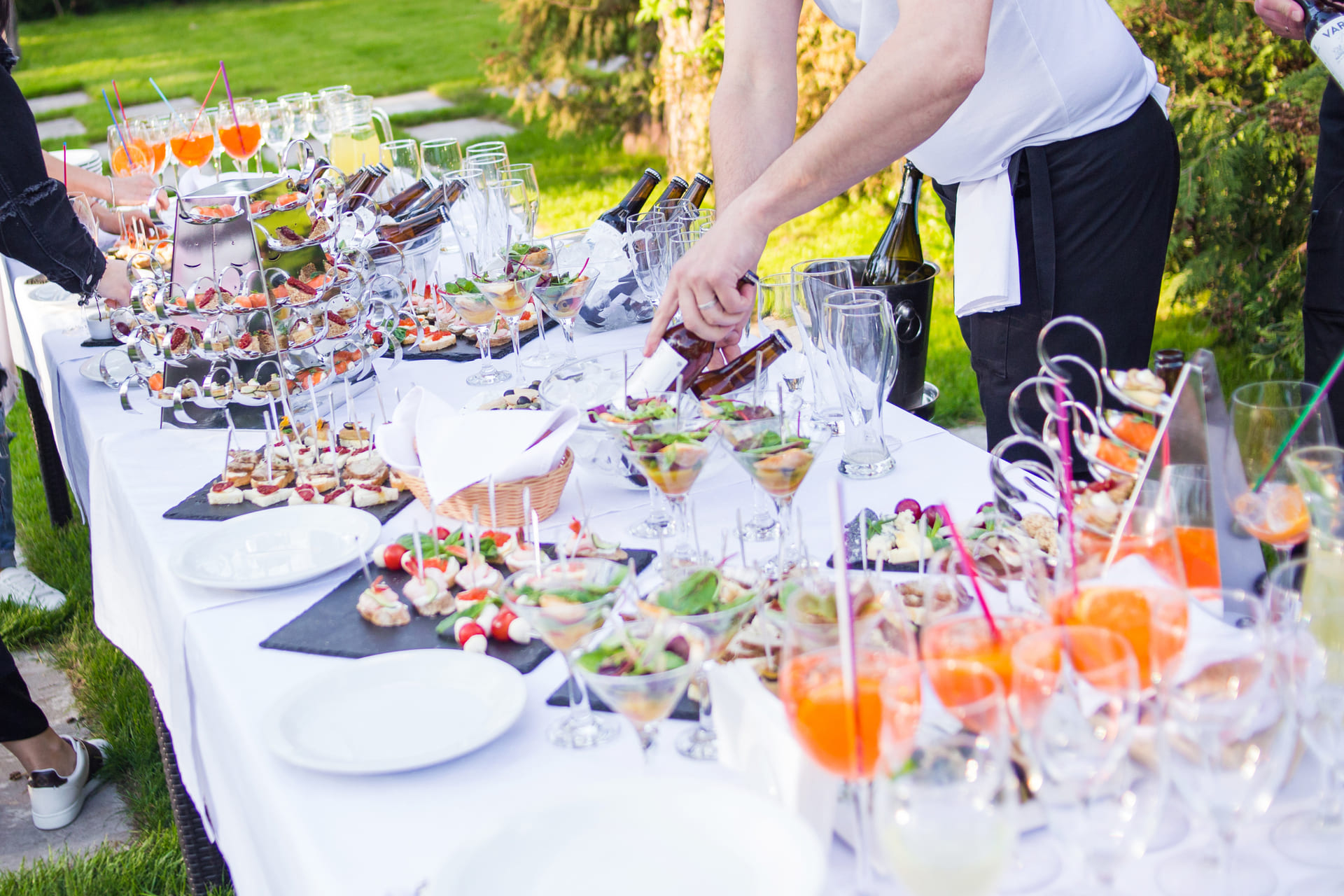 tavoli uniti e posizionati in giardino con buffet di cibo e bevande. Attorno ai tavoli si intravedono degli invitati e un cameriere per il servizio di catering a domicilio